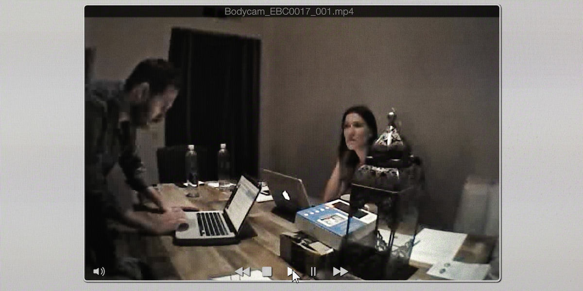 Two people working on laptops at a wooden table in a dimly lit room with water bottles, papers, and a decorative lantern. The scene appears on a computer screen as a paused video.