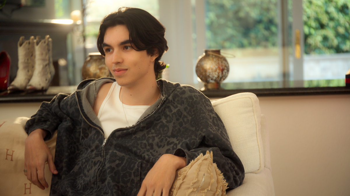 Young person in a patterned hoodie sits on a cream sofa in a bright, stylish living room with a pillow and decorative vases on a counter, with boots in the background and large windows showing greenery outside.