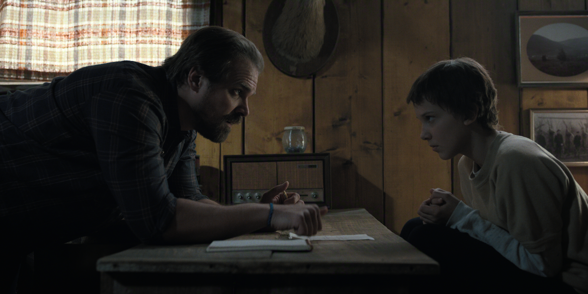A man and a young girl sit across from each other at a wooden table in a dim, rustic room, having an intense conversation.