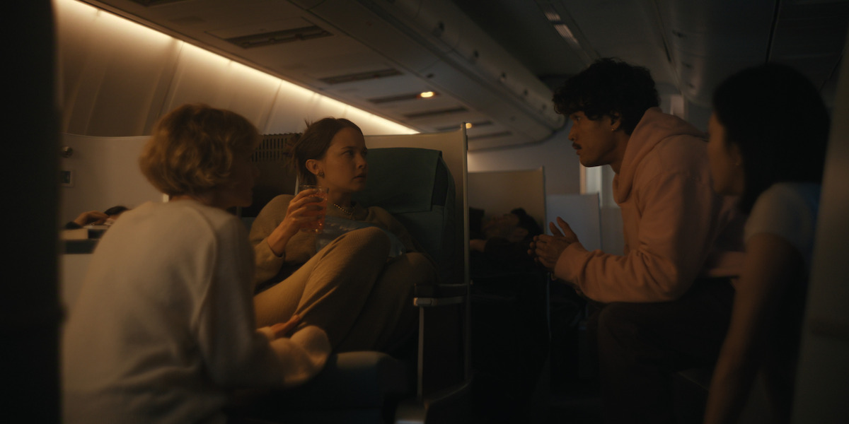 Four people talking in the dimly lit cabin of an airplane, sitting in business class seats with subdued lighting, engaged in a serious conversation at night during a flight.
