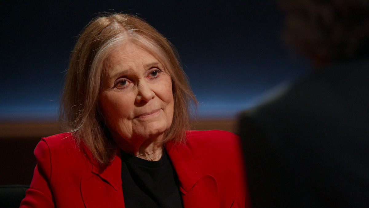 Older woman with shoulder-length hair wearing a red blazer sits indoors under studio lighting, looking attentively at another person whose back is partially visible in the foreground.
