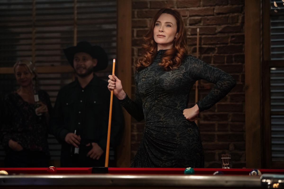 Woman in patterned dress holding pool cue stands confidently by pool table in dimly lit bar with brick wall; two people and drinks in the background.
