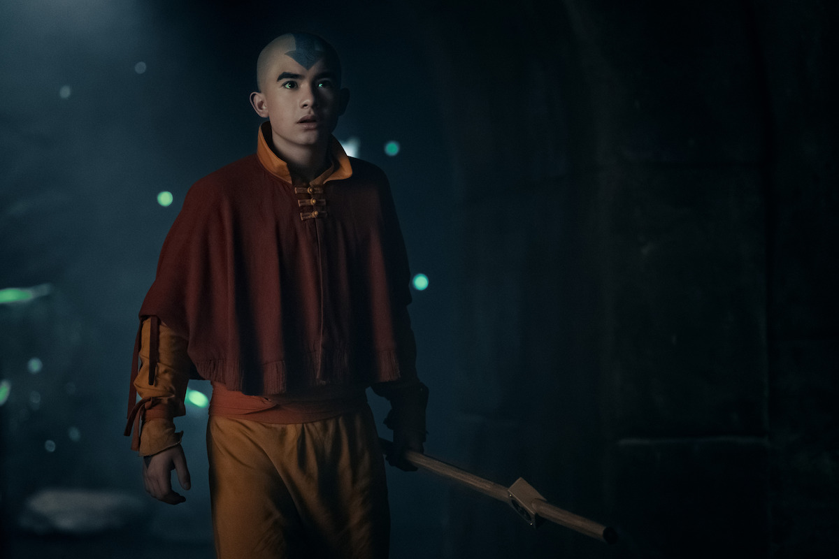 Gordon Cormier as Aang with a blue arrow tattoo on his head, wearing an orange and yellow robe, stands in a dimly lit, mysterious environment holding a wooden staff, looking alert and focused.