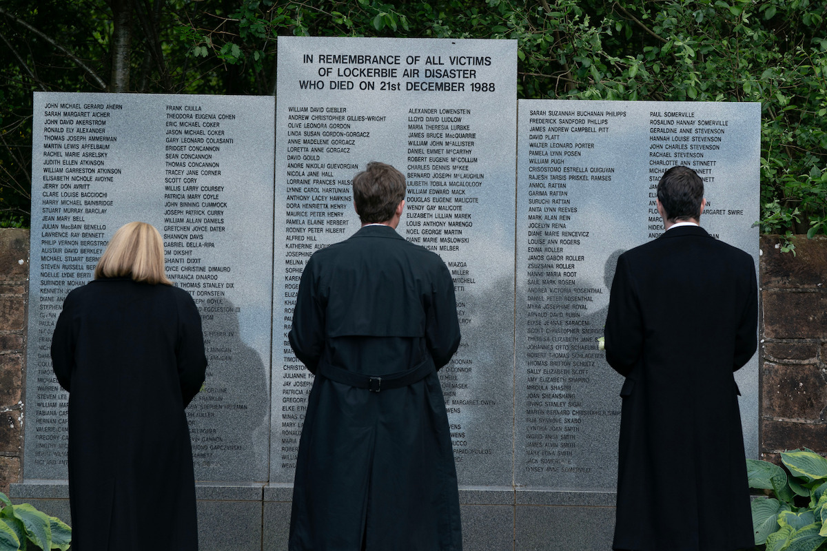 Three people in black coats stand solemnly before a stone memorial engraved with names, honoring victims of the 1988 Lockerbie air disaster, surrounded by greenery.