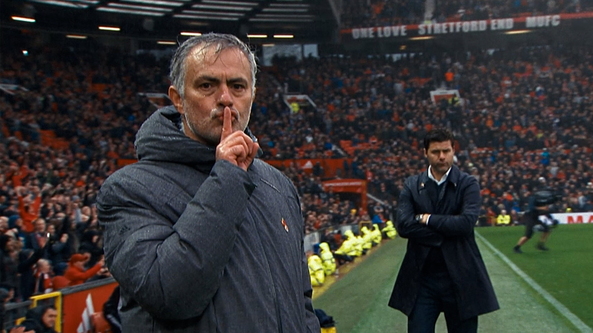 A football manager gestures to the crowd to be quiet during a match at a crowded stadium, with another manager standing in the background, illustrating tension and focus on the sideline during a football game.
