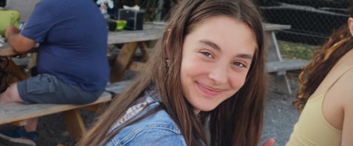 Smiling girl with long brown hair in a casual outdoor setting, sitting at a wooden picnic table with other people and a fence in the background.