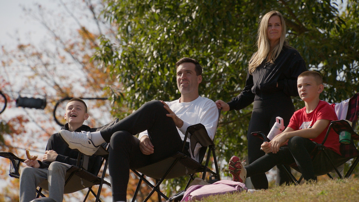 A family of four, two adults and two kids, sit outdoors on folding chairs on a hillside, enjoying a sunny day with trees and autumn foliage in the background.