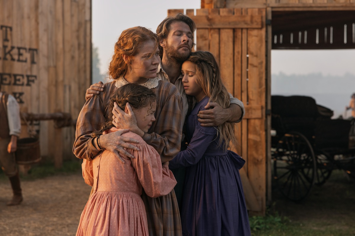A family of four in historical clothing stands hugging outside wooden barn buildings, looking concerned. The background includes wagon wheels, dirt ground, and faded signage, suggesting a rural or frontier setting in the past.