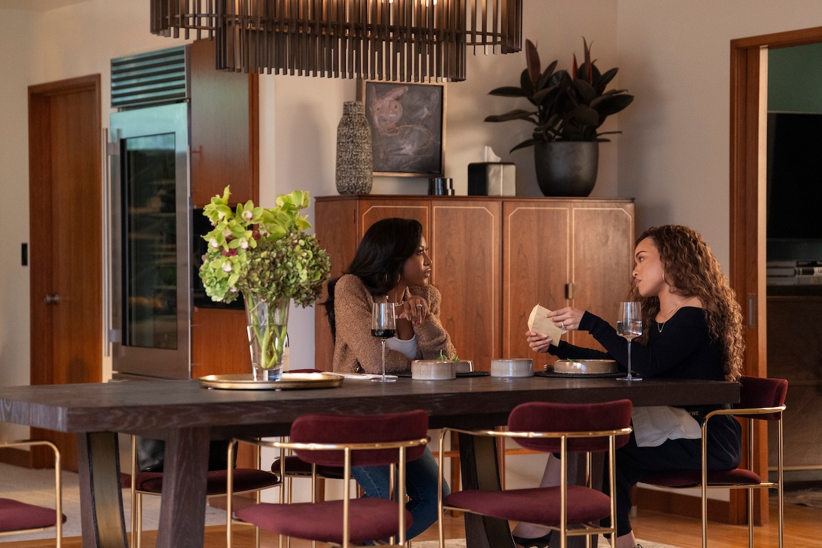 Two women sit at a modern dining table having a conversation over wine and food in a warmly lit, contemporary kitchen with wooden cabinets, decorative plants, and a vase of flowers on the table.