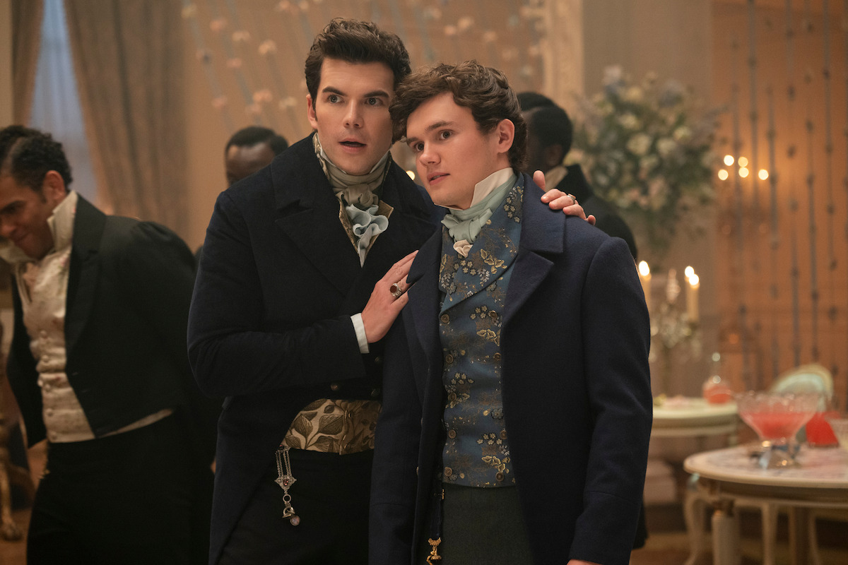 Two young men in formal Regency-era attire stand together at an elegant indoor ball, one with his arm around the other, with guests, candles, flowers, and drinks visible in the ornate background.