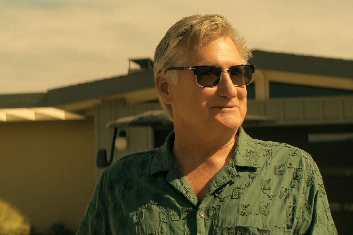 Bill Pullman as Jack in sunglasses and a green shirt standing outside in sunny weather, with a modern house and blue sky in the background.