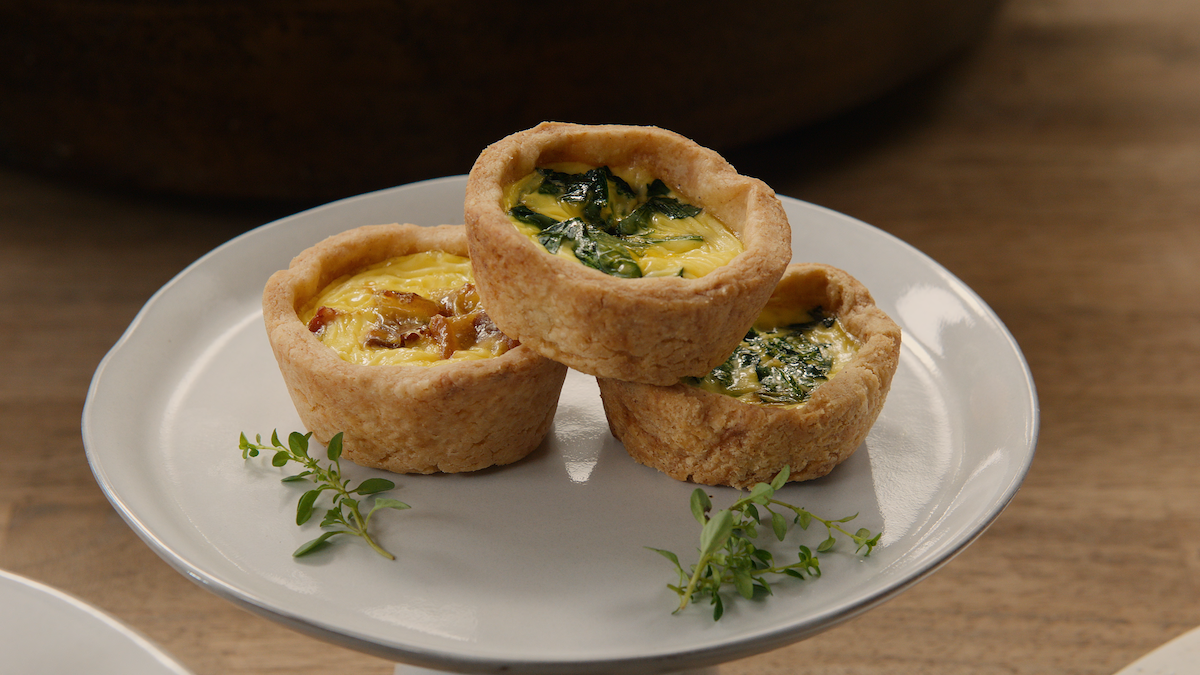 Three mini quiches with golden crusts filled with vegetables and herbs are arranged on a white plate with sprigs of fresh thyme, set on a wooden table, creating a cozy and inviting atmosphere.