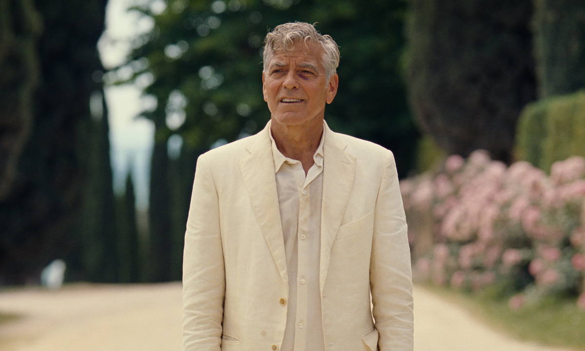 Older man in a light suit stands on a sunlit, tree-lined path with pink flowers in the background, appearing thoughtful and relaxed in a serene, lush outdoor setting.