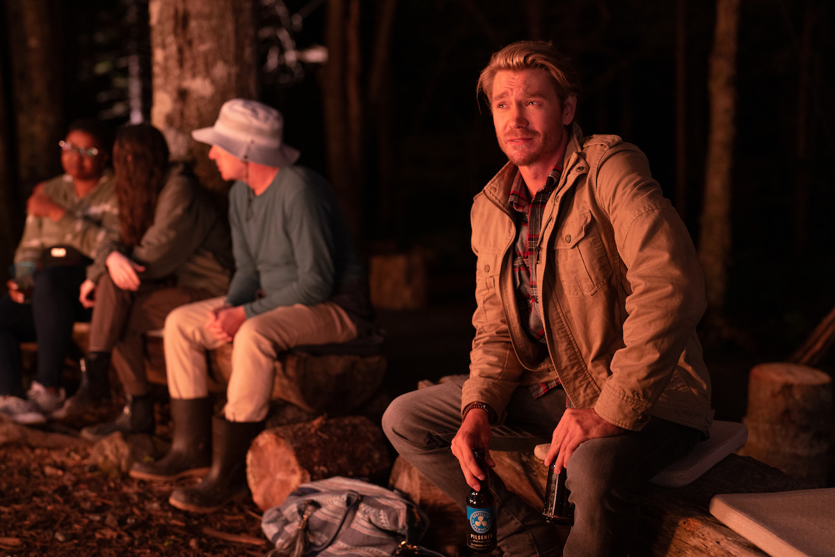 In a picture of 'Sullivan's intersection' Chad Michael Murray holds a bear as he wears a jacket while sitting on a bench in front of the glow of a fire.