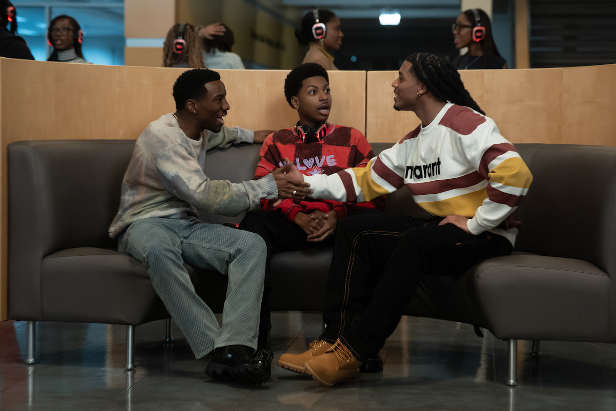Three young men sitting on modern couches, talking animatedly in a public indoor space; two are shaking hands while the third looks surprised. People in the background wear headphones, suggesting a silent event or party setting.