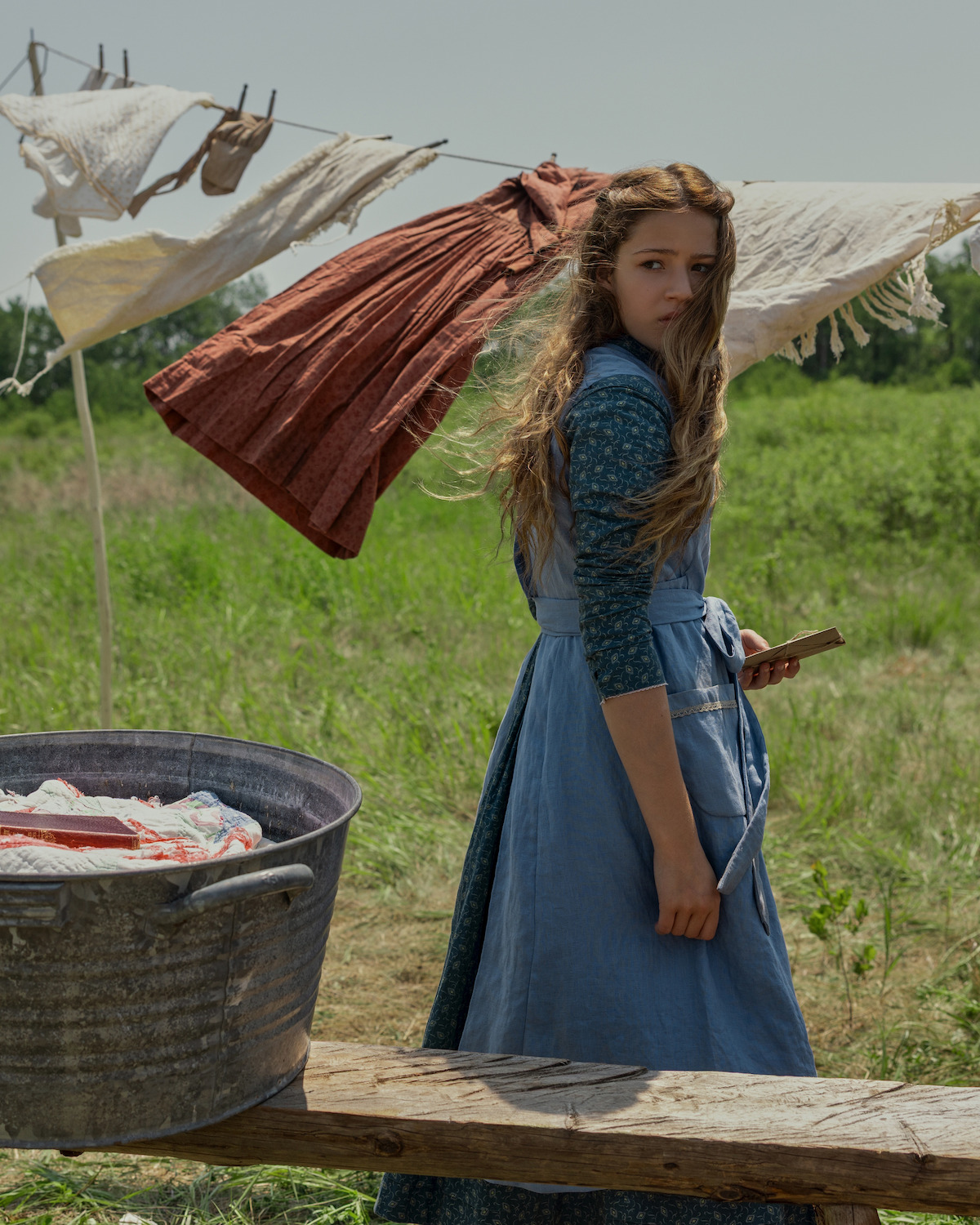 Young girl in old-fashioned blue dress holding a book, standing outdoors beside a wooden bench and metal washtub with laundry. Clothes hang on a line blowing in the wind, with green grass field and trees in the background.