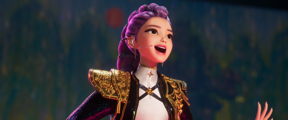 Animated female character with purple hair and gold-embellished jacket singing on stage, wearing a headset microphone. Blurred dark background suggests a theatrical or concert performance setting.