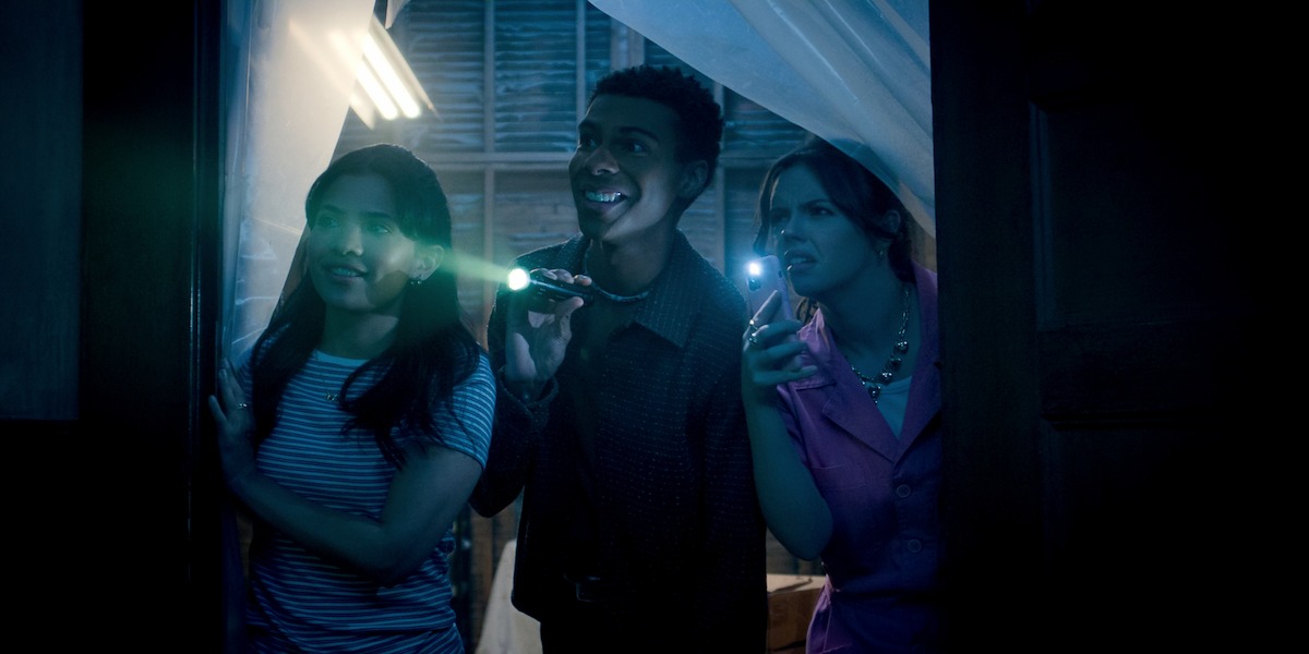 Three young adults exploring a dimly lit room with flashlights, appearing curious and excited, standing near a window with sheer curtains and wooden walls visible in the background.