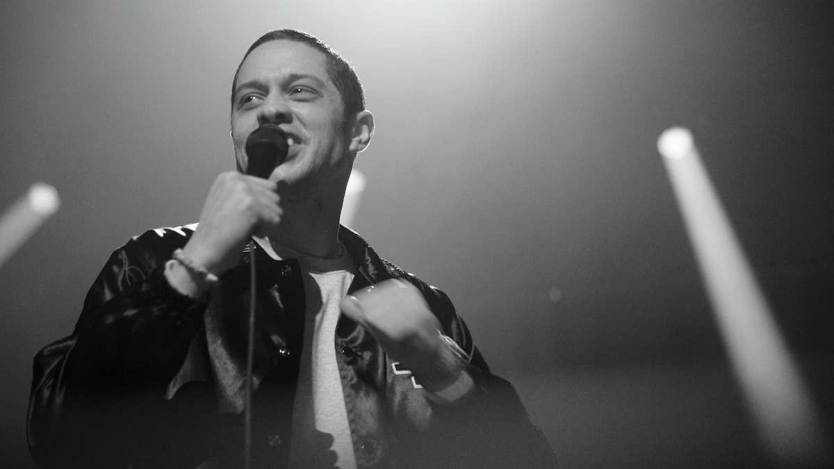 Un hombre que sostiene un micrófono actúa en el escenario en un ambiente oscuro y lleno de humo con luces y haces dramáticos, pareciendo estar comprometido con la audiencia.