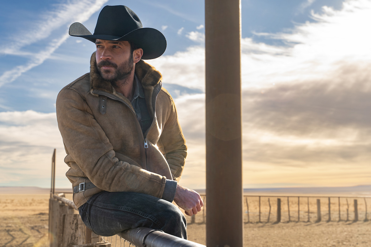 Man wearing cowboy hat and jacket sits on a wooden fence in a wide open rural landscape at sunset, with a cloudy sky and distant mountains, evoking a calm, rugged western atmosphere.