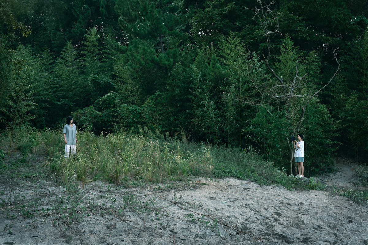 Two people standing apart in a grassy, sandy clearing at the edge of a dense forest, one near tall grass and the other by a small tree, creating a quiet and contemplative mood in a natural outdoor setting.