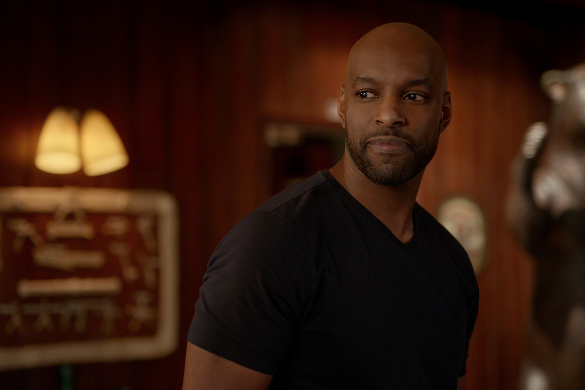 A man with a beard and shaved head wearing a dark t-shirt stands indoors in a warmly lit room with wooden walls, a lamp, and a bear statue in the background, giving a thoughtful or contemplative expression.