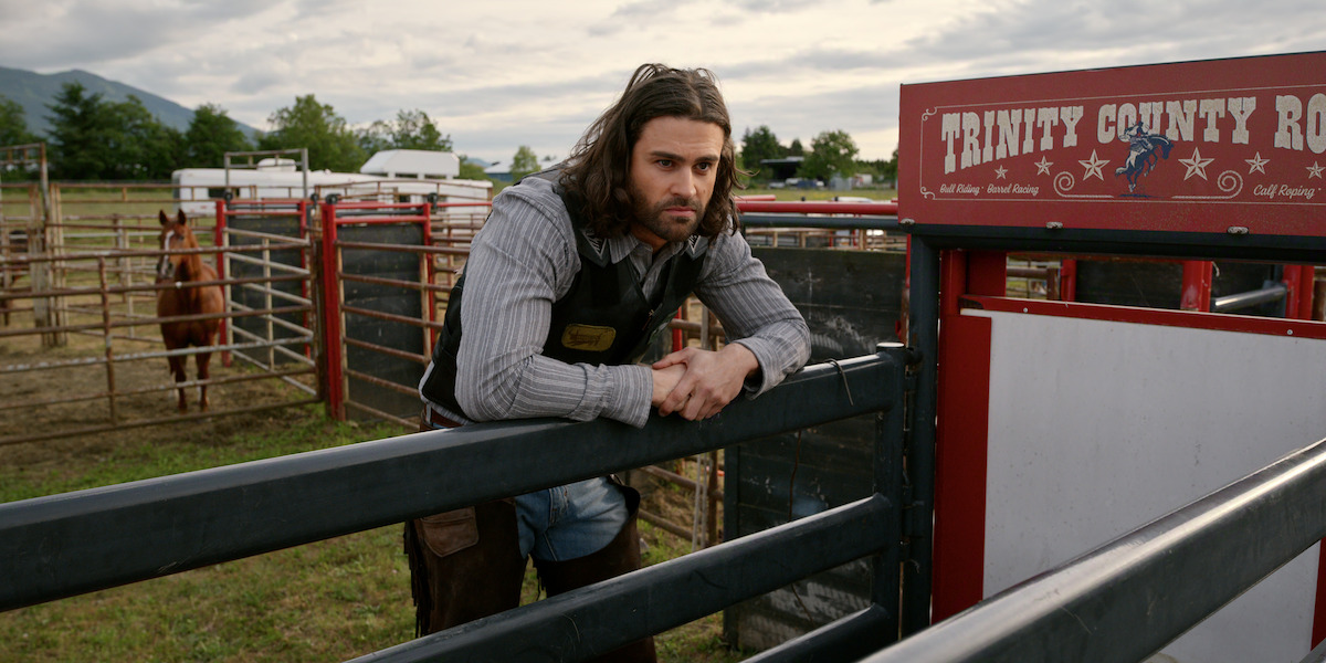 A man with long hair and a beard, dressed in rodeo gear, leans on a metal gate in a rural outdoor rodeo arena with a horse nearby and trees in the background under a cloudy sky.