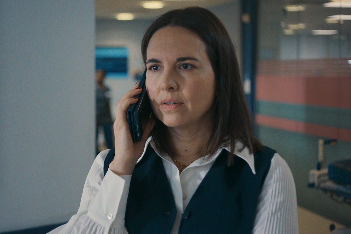 Una mujer de cabello castaño, vestida con una camisa blanca y un chaleco oscuro, habla por un teléfono celular en una oficina moderna o en el pasillo de un hospital, luciendo seria.