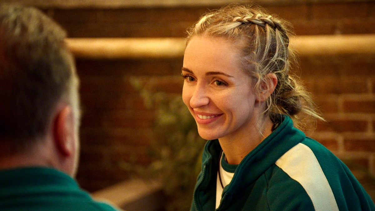 A woman in a green sports jacket smiles warmly at someone across from her in a cozy indoor setting with brick walls, creating a friendly and relaxed mood.
