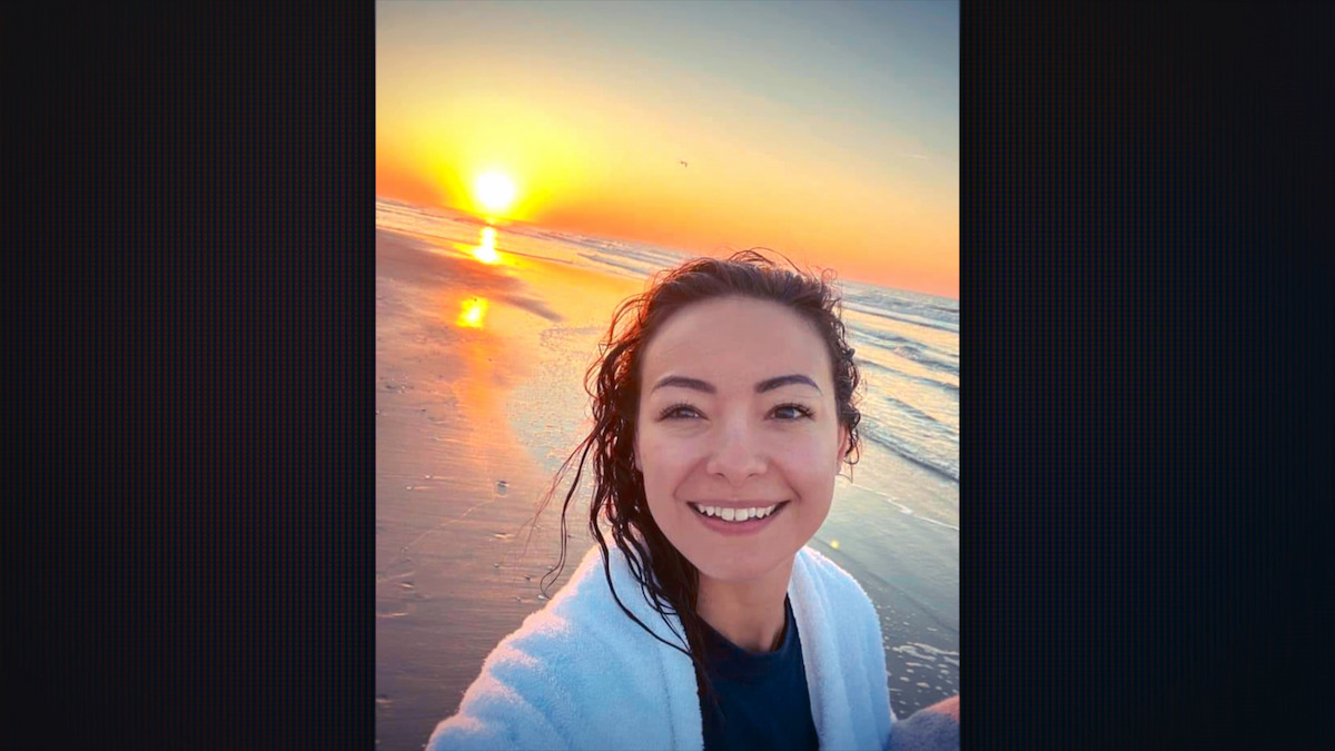Smiling woman taking a selfie on a beach at sunrise, wearing a white towel, with wet hair, the sun low over the ocean, and waves gently rolling onto the shore under a colorful sky.