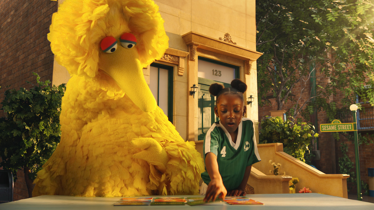 Big Bird and a young girl, both at a table with cards, sit outdoors in front of a brownstone building on Sesame Street, with greenery and street signs in the background.