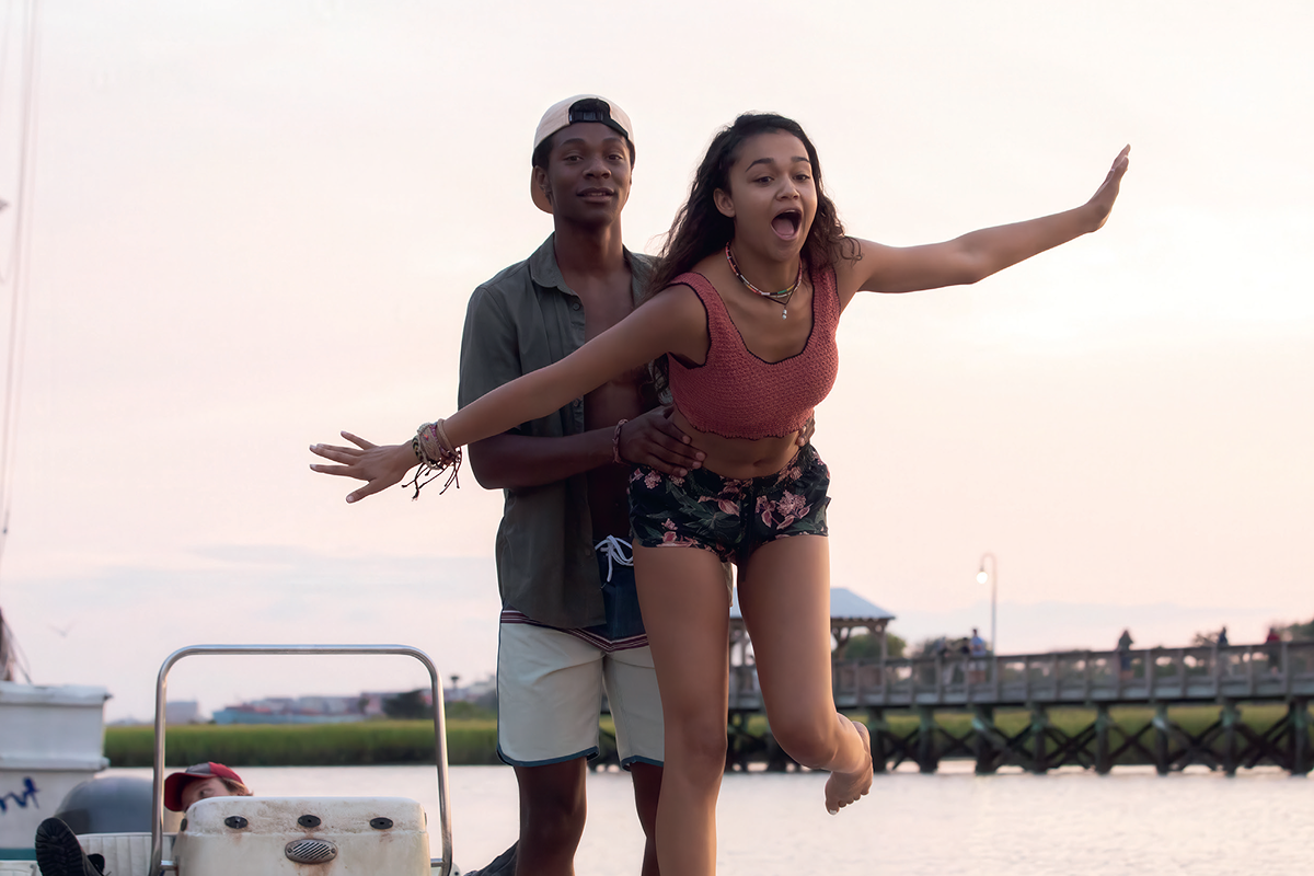 Two young people on a boat at sunset, one playfully leaning forward with arms outstretched while the other holds on from behind, with a wooden dock and water in the background.