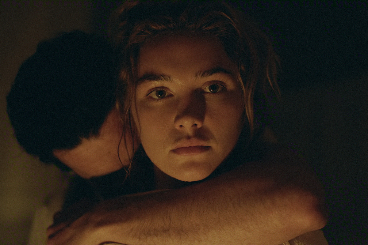 A woman with a serious expression is embraced by a man in a dimly lit, intimate indoor setting, highlighting her face and emotions while most of his face is hidden in shadow.