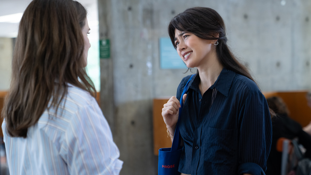 Two women are having a friendly conversation indoors in a modern setting with concrete walls, natural light, and other people in the background.