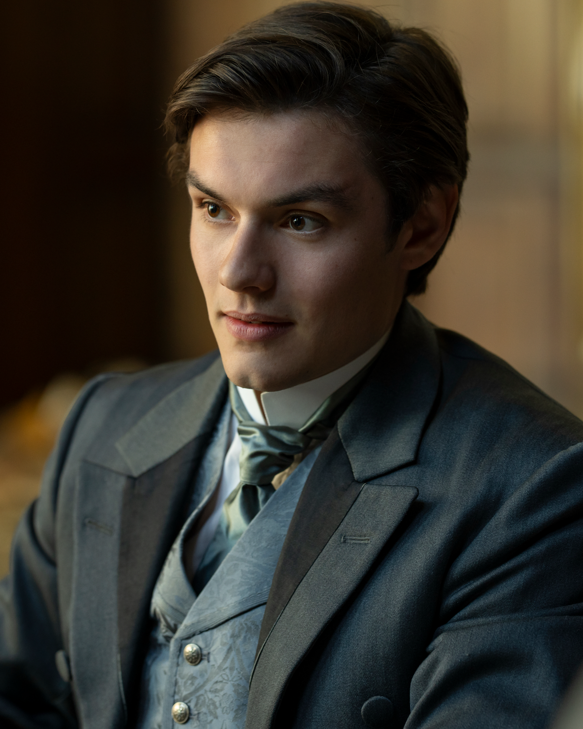 Young man in a Victorian-style suit with vest and cravat, seated indoors in a warmly lit, wooden-paneled room, looking attentively to the side.