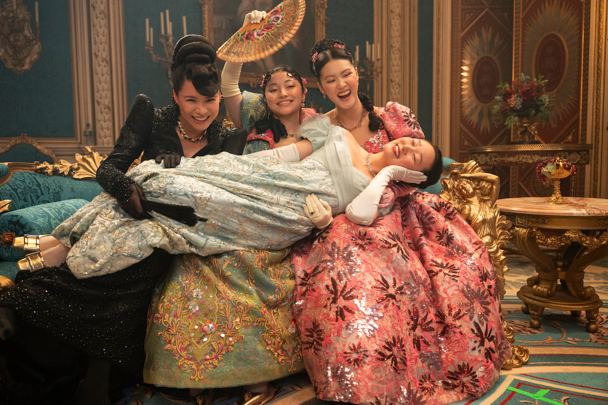 Four women in elegant, colorful period costumes laugh together in a lavish, gold-trimmed room, with three sitting on a couch holding another woman lying across their laps, creating a joyful and playful atmosphere.