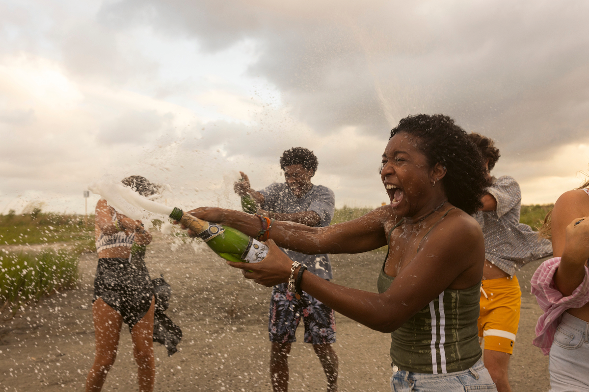 A joyful woman in a green tank top sprays champagne outdoors with friends on a cloudy day, celebrating together on a rural road with grass and open sky in the background.