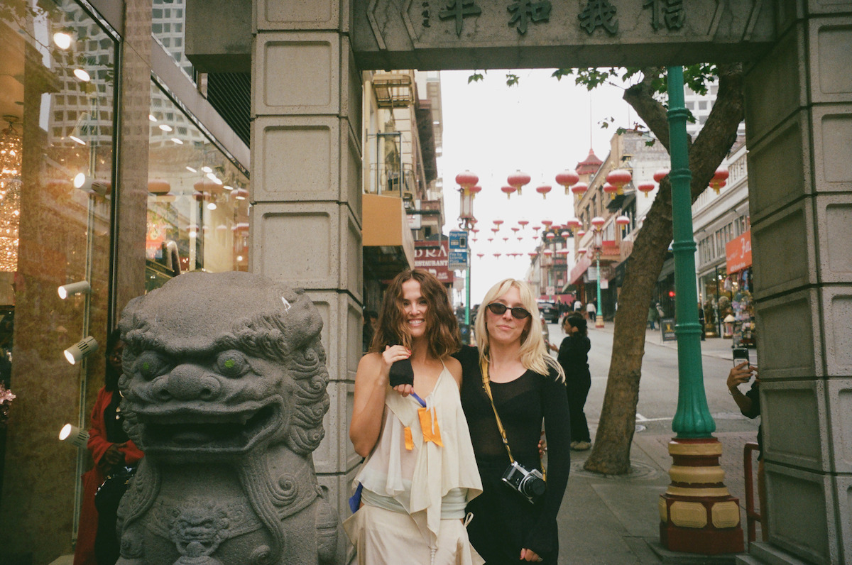Two women pose next to a stone lion statue under an ornate gate in a vibrant Chinatown street, with hanging red lanterns, storefronts, and people in the background on a cloudy day.