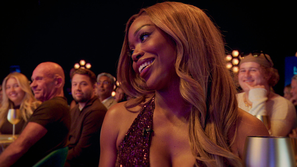 Smiling woman with long hair in a sparkly dress sits at a table in a lively, dimly lit event with other people in the background, warmly lit by decorative lights.