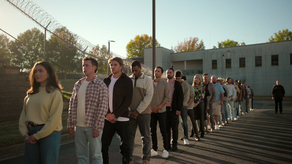 A group of people standing in a line outdoors next to a barbed wire fence and a large concrete building, possibly at a prison or secured facility, with trees and sunlight in the background.