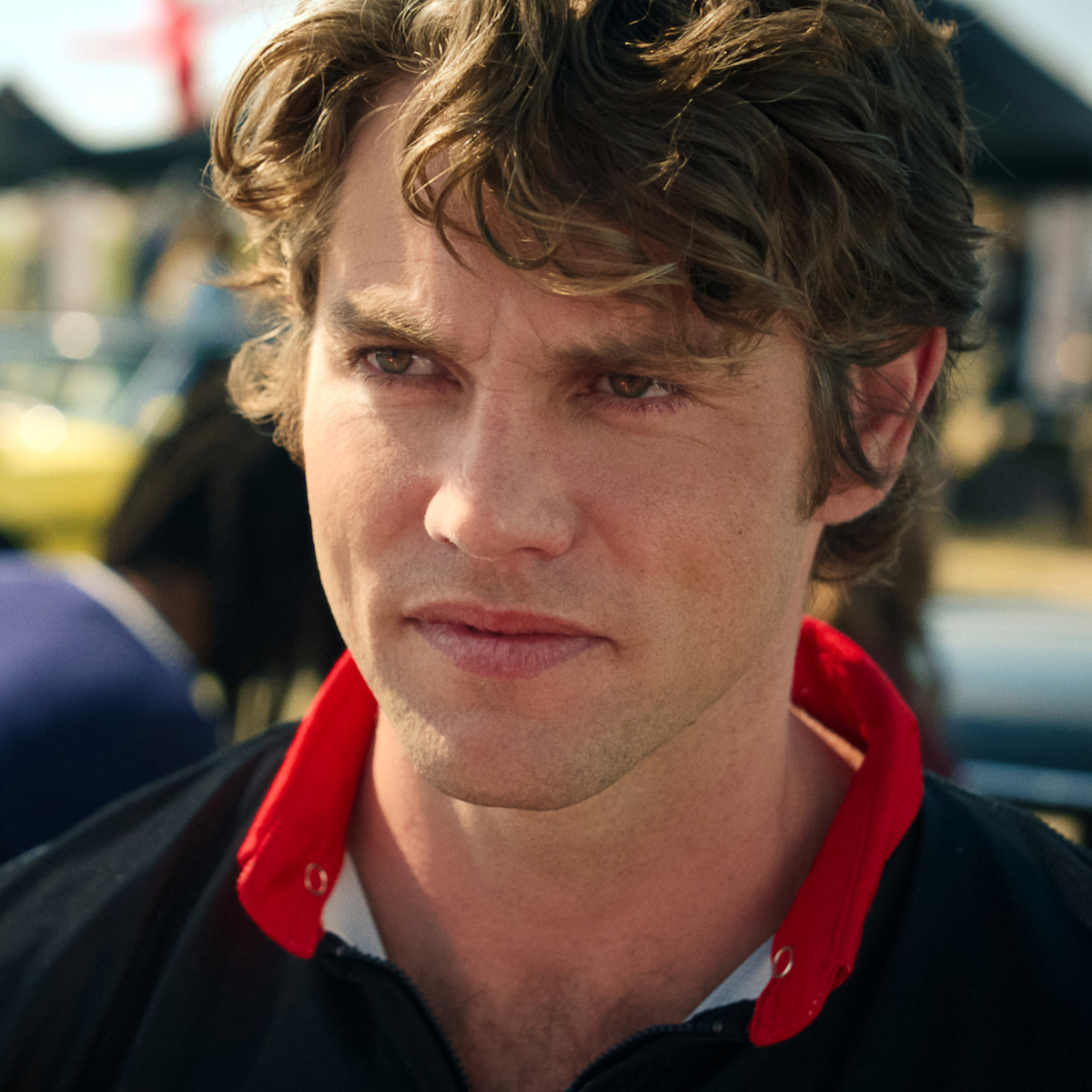 A man with wavy brown hair in a black and red collared shirt stands outdoors, likely at a car show or race, with blurred cars, people, and tents in the sunny background.