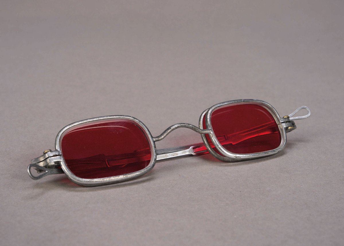 Vintage silver-framed eyeglasses with red-tinted lenses resting on a plain, neutral gray surface, minimalist backdrop, calm and subdued mood.