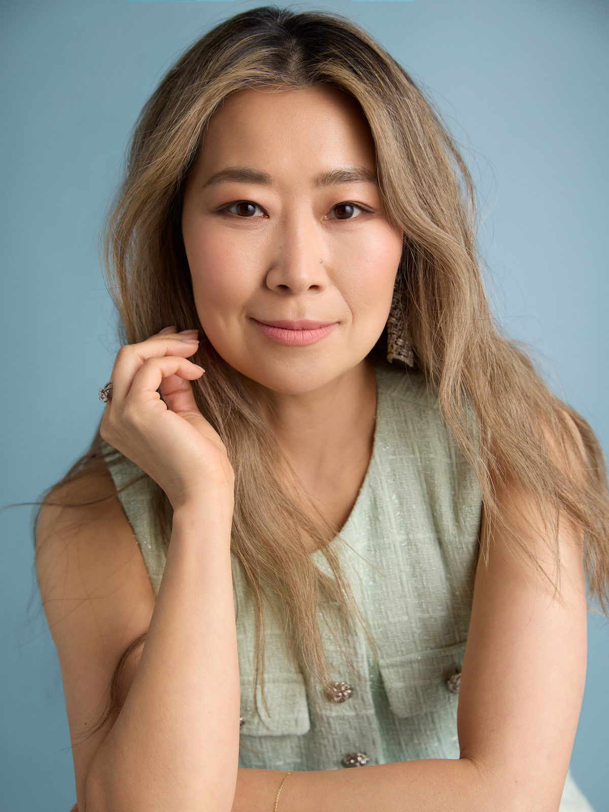Maggie Kang with long, light brown hair poses in a sleeveless light green top against a plain light blue background, gazing softly at the camera with a relaxed and thoughtful expression.