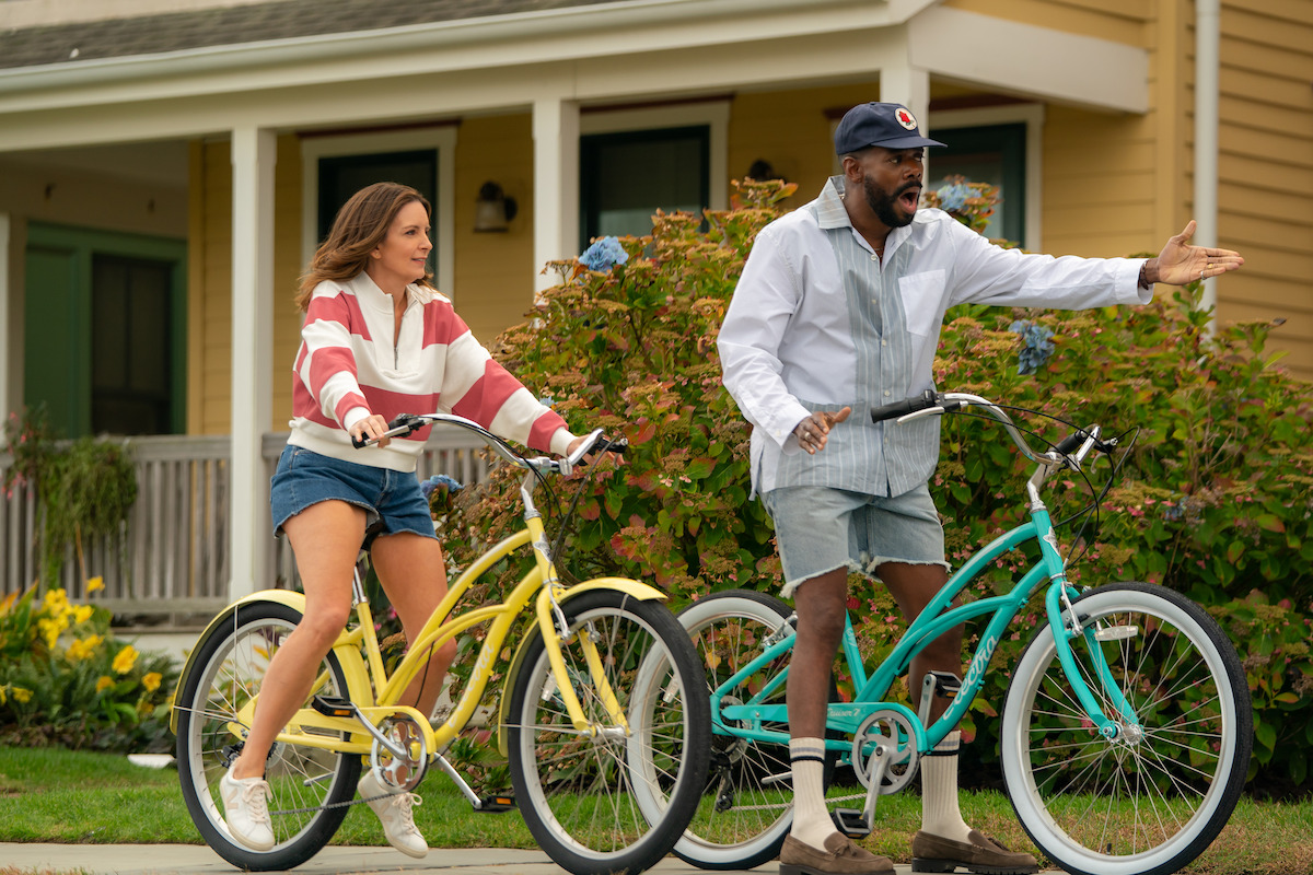 Two adults ride bicycles on a suburban street lined with greenery and yellow houses; one is gesturing and talking animatedly while the other listens, both wearing casual summer clothes.