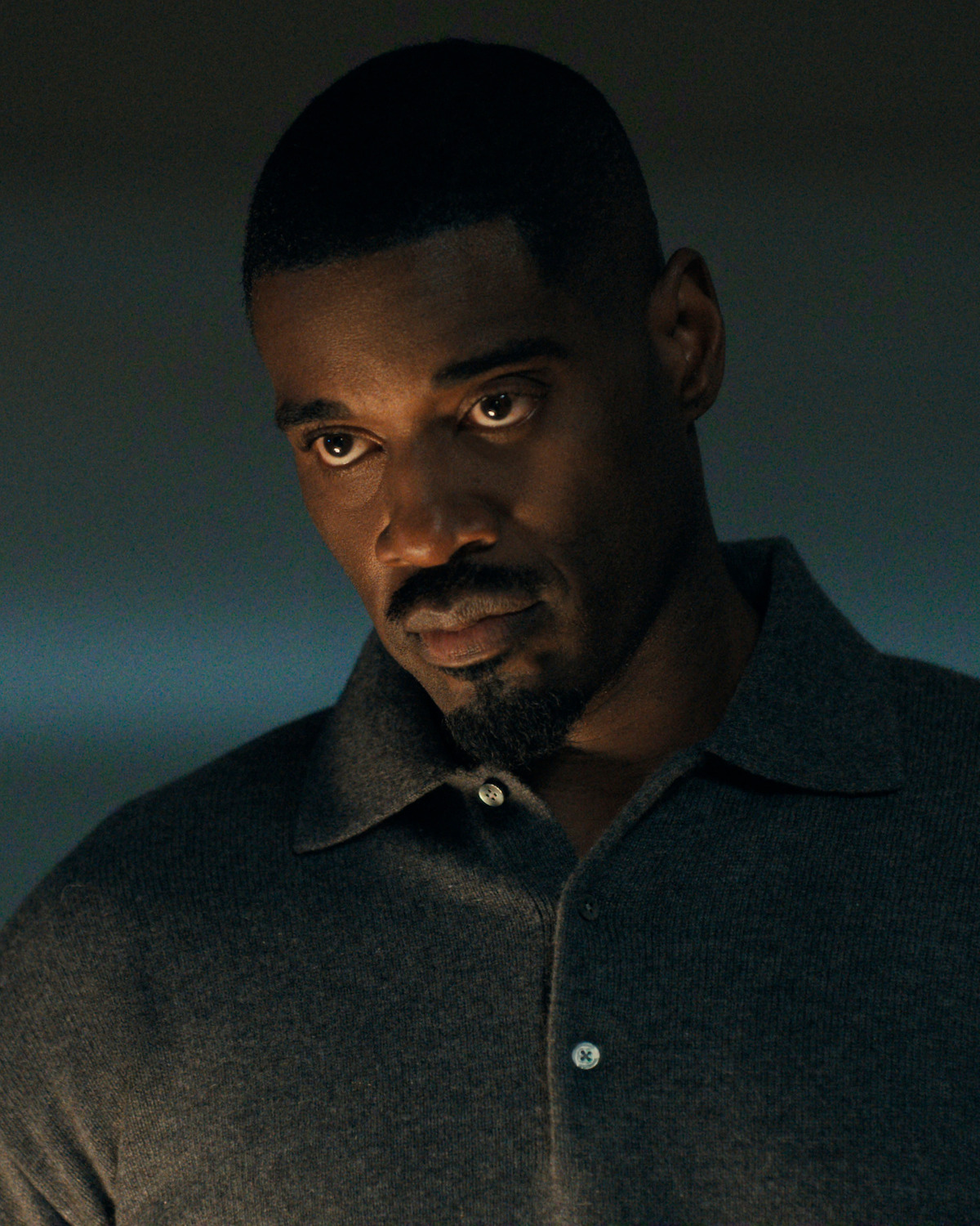 Man with short hair and goatee wearing a dark buttoned shirt in a dimly lit indoor setting, looking contemplative.