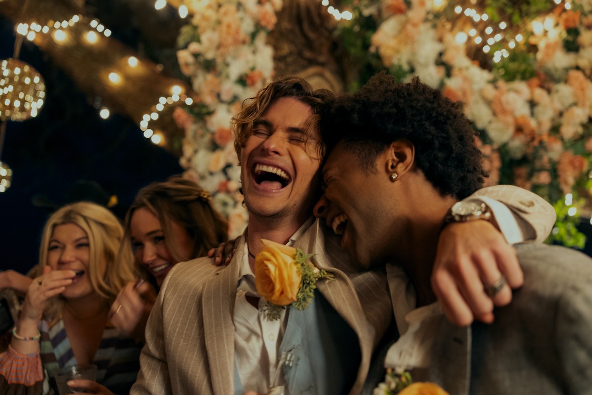 Two friends in suits laughing and embracing at a festive outdoor event with string lights and floral decorations, surrounded by smiling guests at night.
