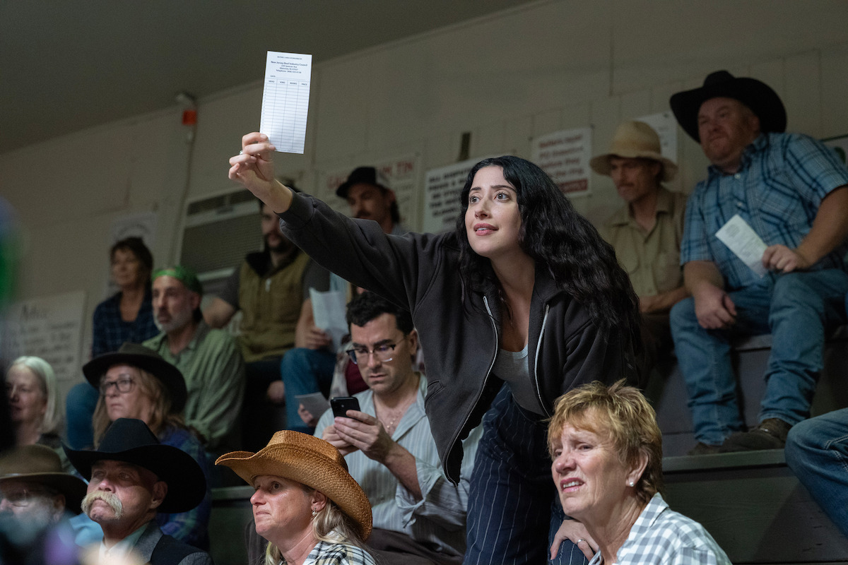 Woman holding up a bidding slip in a crowded indoor auction, surrounded by people in casual and western attire sitting on bleachers, focused on the auction event.