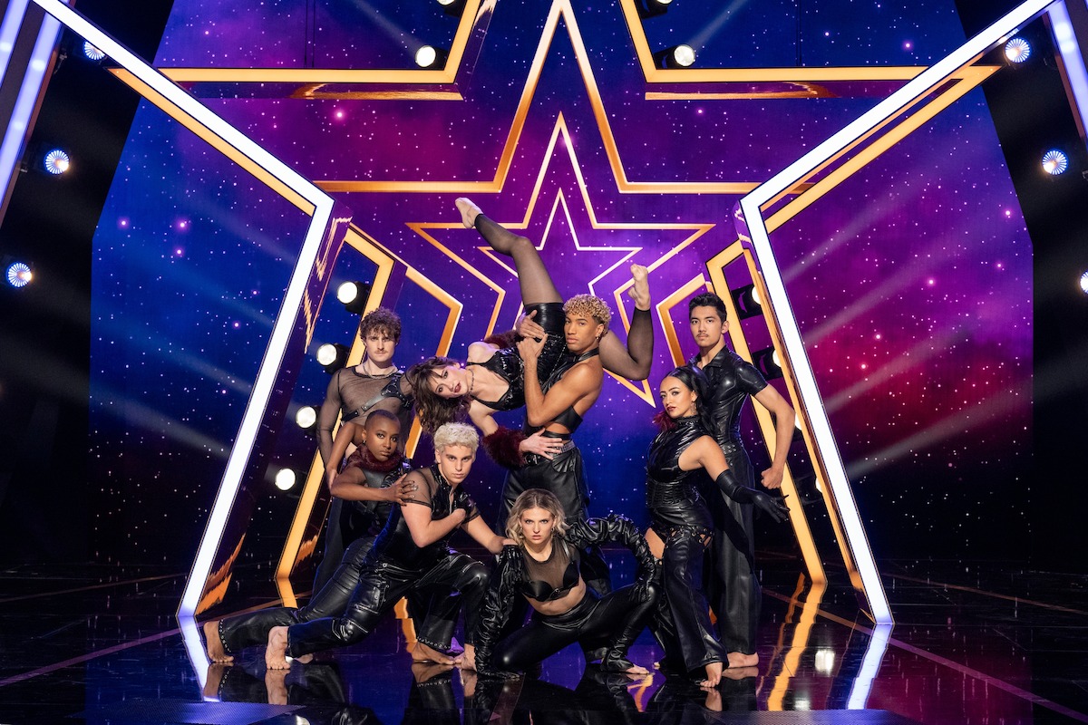 Movement 55 posa dramáticamente en un escenario brillante con un fondo de estrellas moradas y luces brillantes, los artistas visten trajes negros coordinados, mostrando una coreografía dinámica y un fuerte trabajo en equipo.