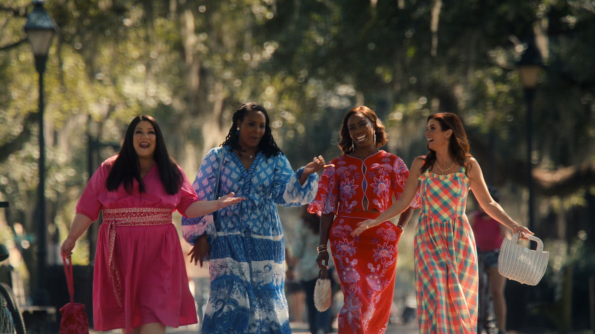 Four women in colorful dresses walk together outdoors along a sunlit, tree-lined path, smiling and talking, with relaxed park surroundings and dappled sunlight in the background.