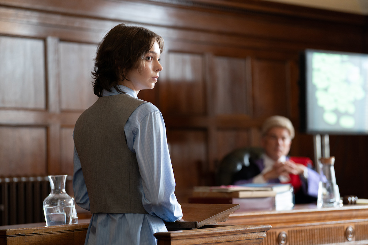 A young person stands at a wooden podium in a courtroom, facing forward, with a judge seated in the background near legal documents, a glass jug, and a blurred computer screen displaying information.